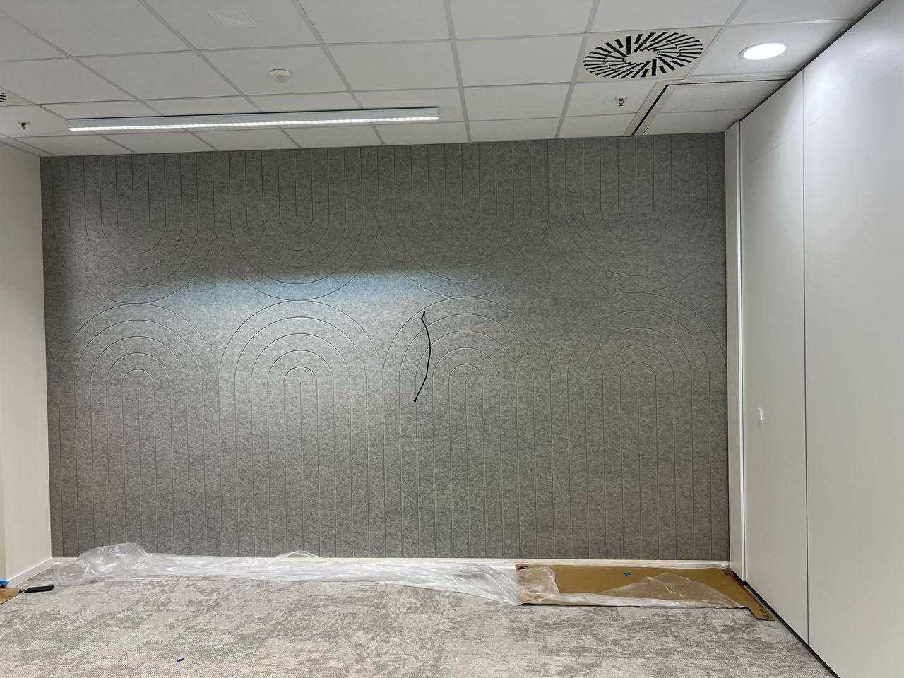 Szara ściana z filcowymi panelami akustycznymi w kształcie łuków, zabezpieczona folią malarską. Widok na fragment podłogi i białą szafę w biurze.