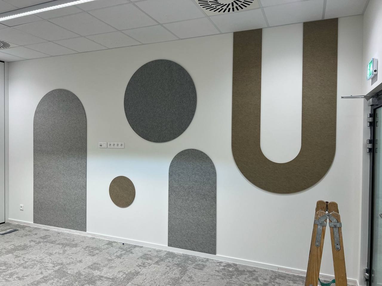 Biuro z dekoracyjnymi panelami ściennymi w kształcie figur geometrycznych (koła i półkola) i litery U. Na podłodze szara wykładzina dywanowa, w rogu drabina.