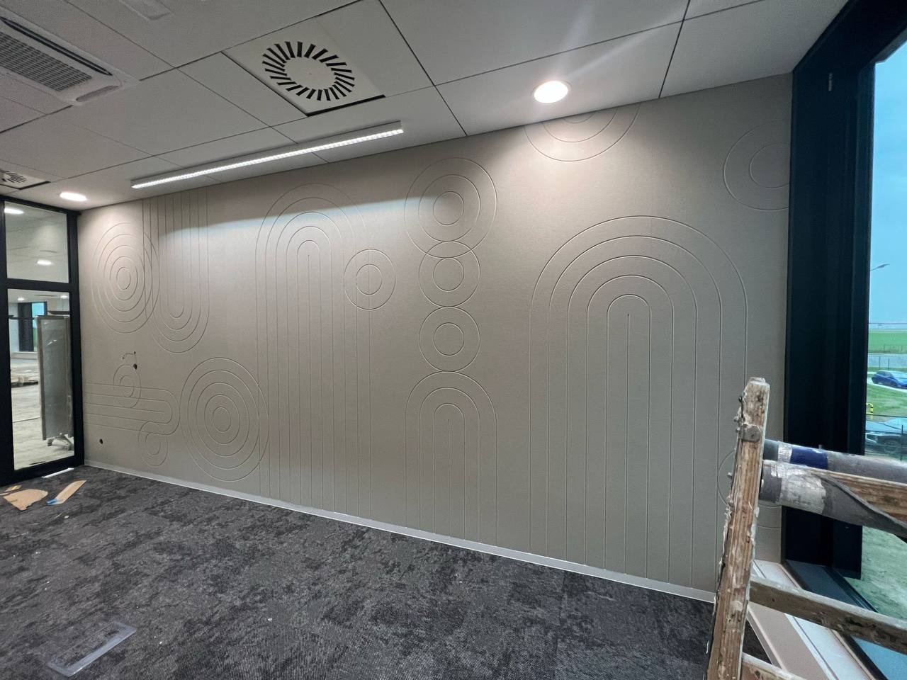 Wykończona ściana z dekoracyjną, geometryczną fakturą w odcieniach szarości. Widoczne elementy drabiny malarskiej i fragment okna. Nowoczesne wnętrze biurowe.