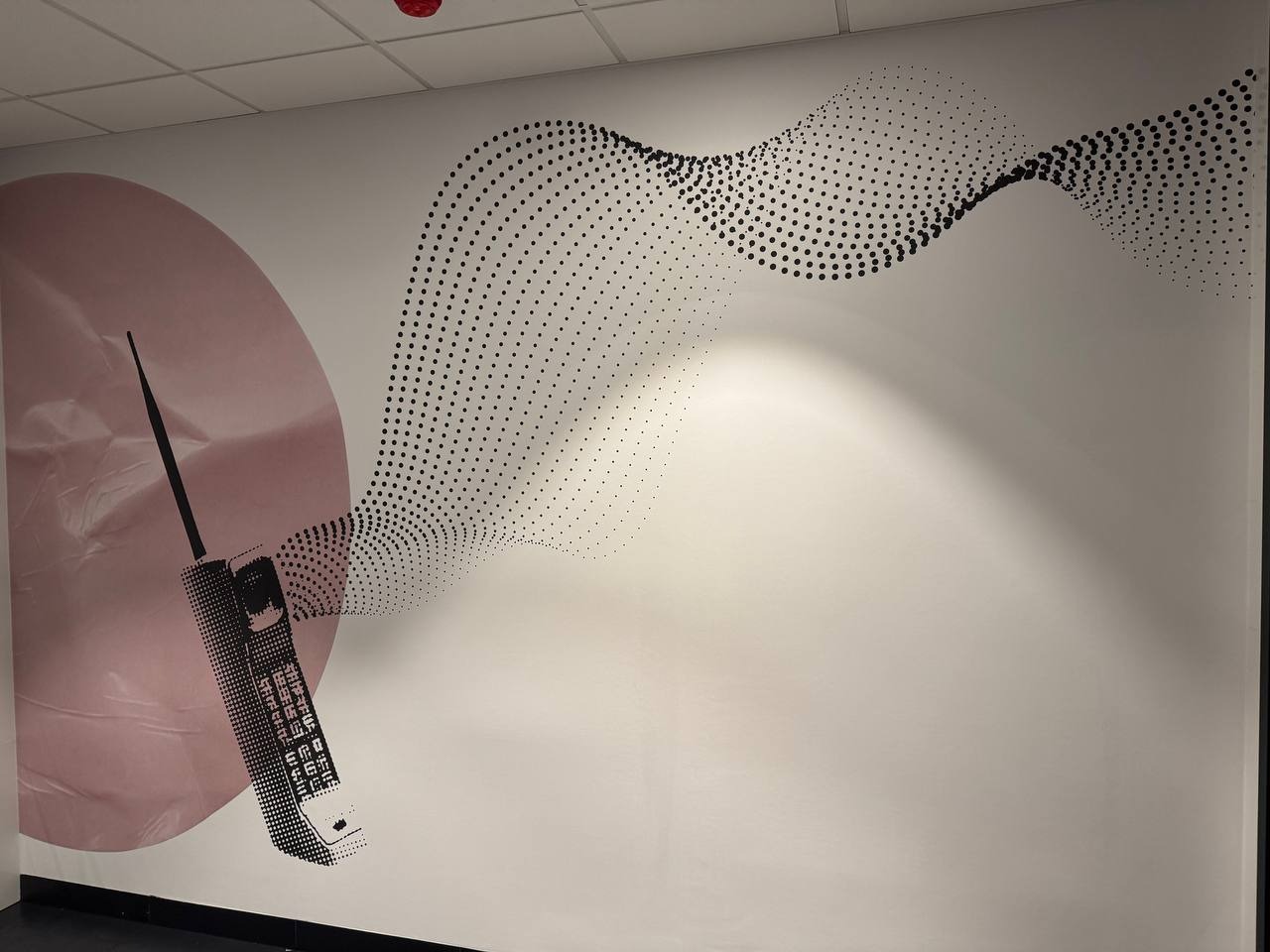 Ściana z tapetą przedstawiającą duży, czarno-biały telefon komórkowy z anteną na tle różowego koła i wzorem z kropek. Oświetlenie z góry.