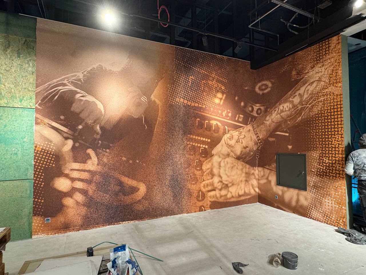 Pokój z fototapetą przedstawiającą DJ-a za konsoletą, w odcieniach brązu. Widoczne narzędzia malarskie i fragment sylwetki osoby w tle.