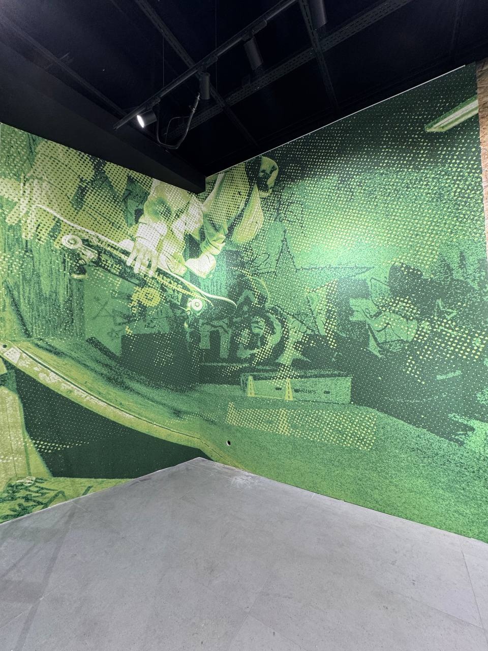 Zielona tapeta z motywem skateparku, przedstawiająca postać na deskorolce. Ujęcie z dołu, widoczny fragment podłogi i sufitu. Nietypowy, młodzieżowy design.