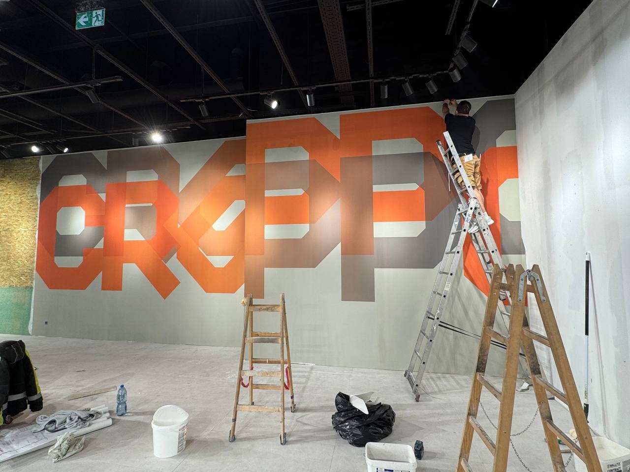 Pracownik na drabinie montuje wielkoformatową tapetę z geometrycznym wzorem w odcieniach pomarańczy i szarości w przestronnym wnętrzu z oświetleniem punktowym.