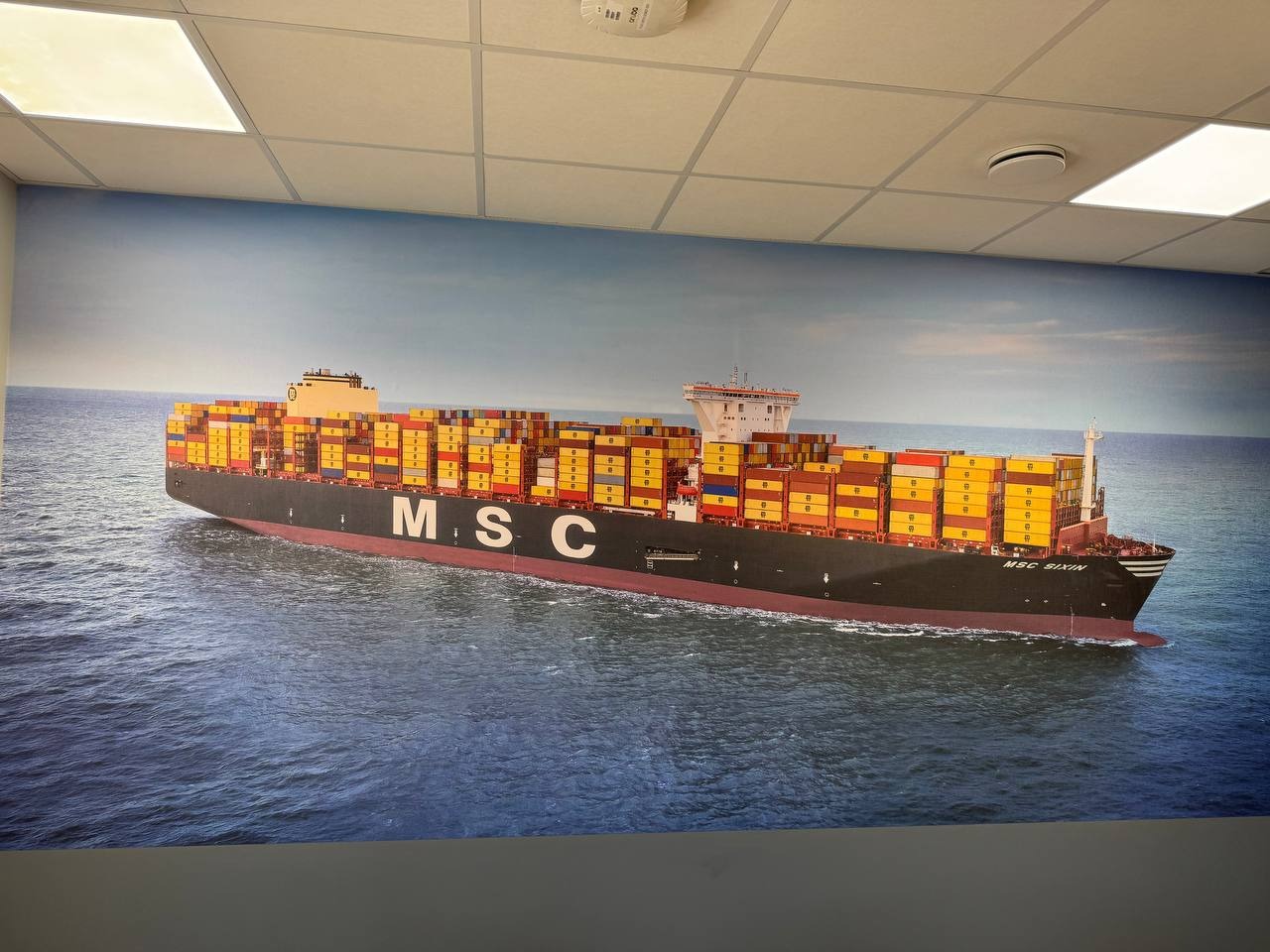 Fototapeta z motywem kontenerowca MSC SIXIN na ścianie biura, widoczna pod sufitem z oświetleniem rastrowym.