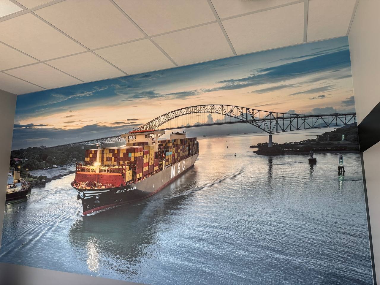 Fototapeta z widokiem kontenerowca przepływającego pod mostem, naklejona na ścianie w pomieszczeniu z kasetonowym sufitem.