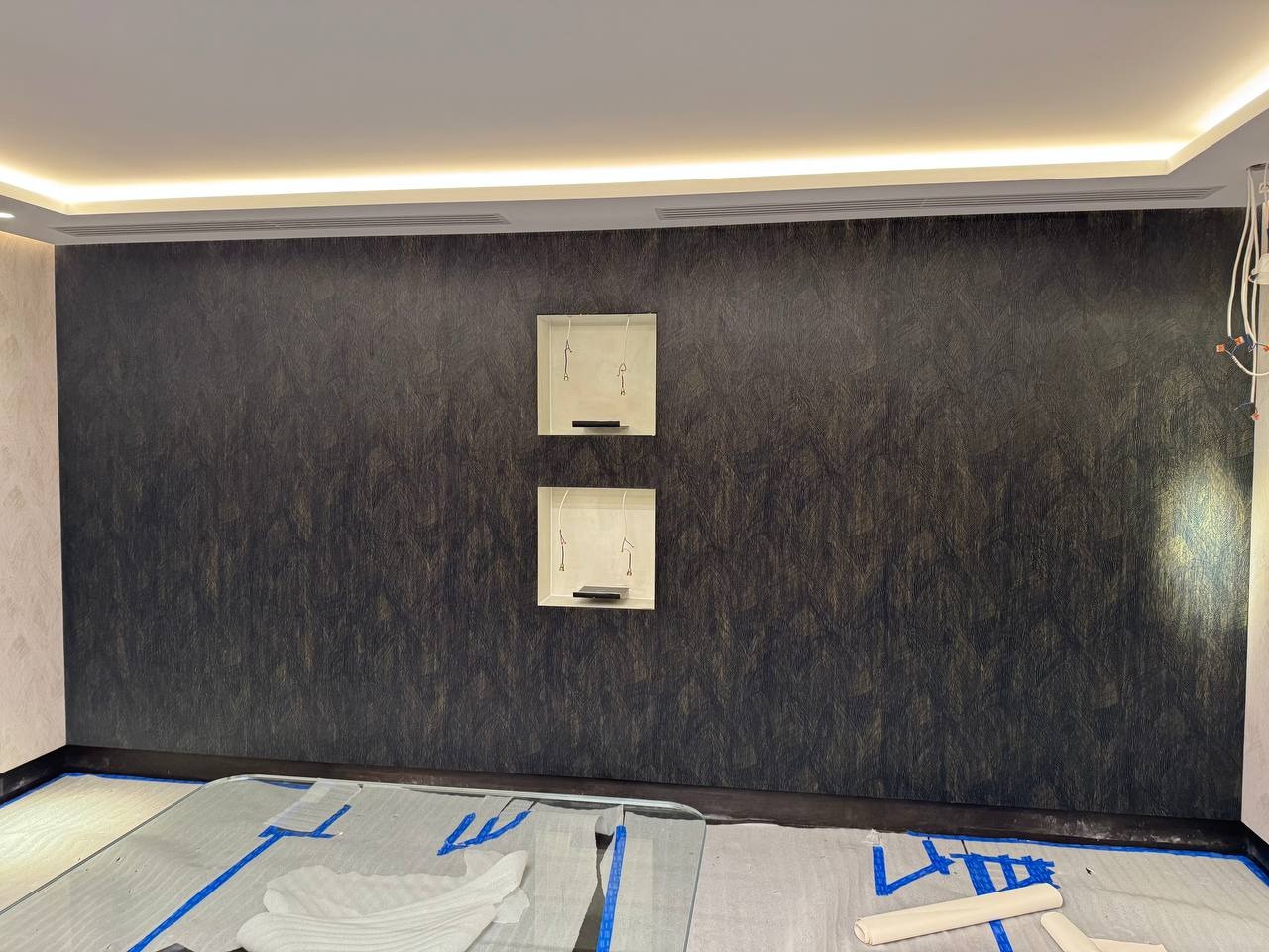 Wykończone ściany z ciemną tapetą o wzorze przypominającym liście w pokoju z zabezpieczoną podłogą i wnękami z oświetleniem punktowym.