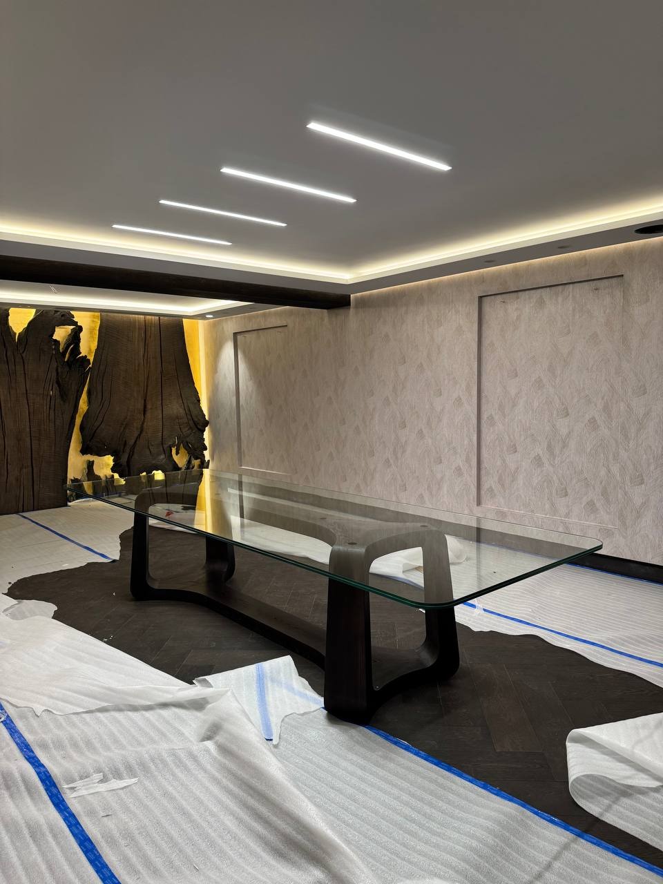 Eleganckie wnętrze z tapetą w neutralnych barwach i nowoczesnym stołem. Widoczne zabezpieczenia podłogi sugerują świeżo zakończone prace wykończeniowe.