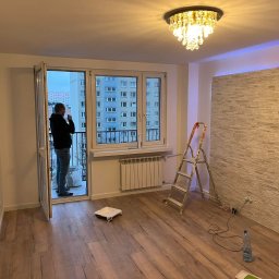 MDM bud - Jasne wnętrze po remoncie: drewnopodobna podłoga, ściana z dekoracyjnej cegły, drabina malarska, otwarte drzwi balkonowe z widokiem na blok. Oświetlenie sufitowe.