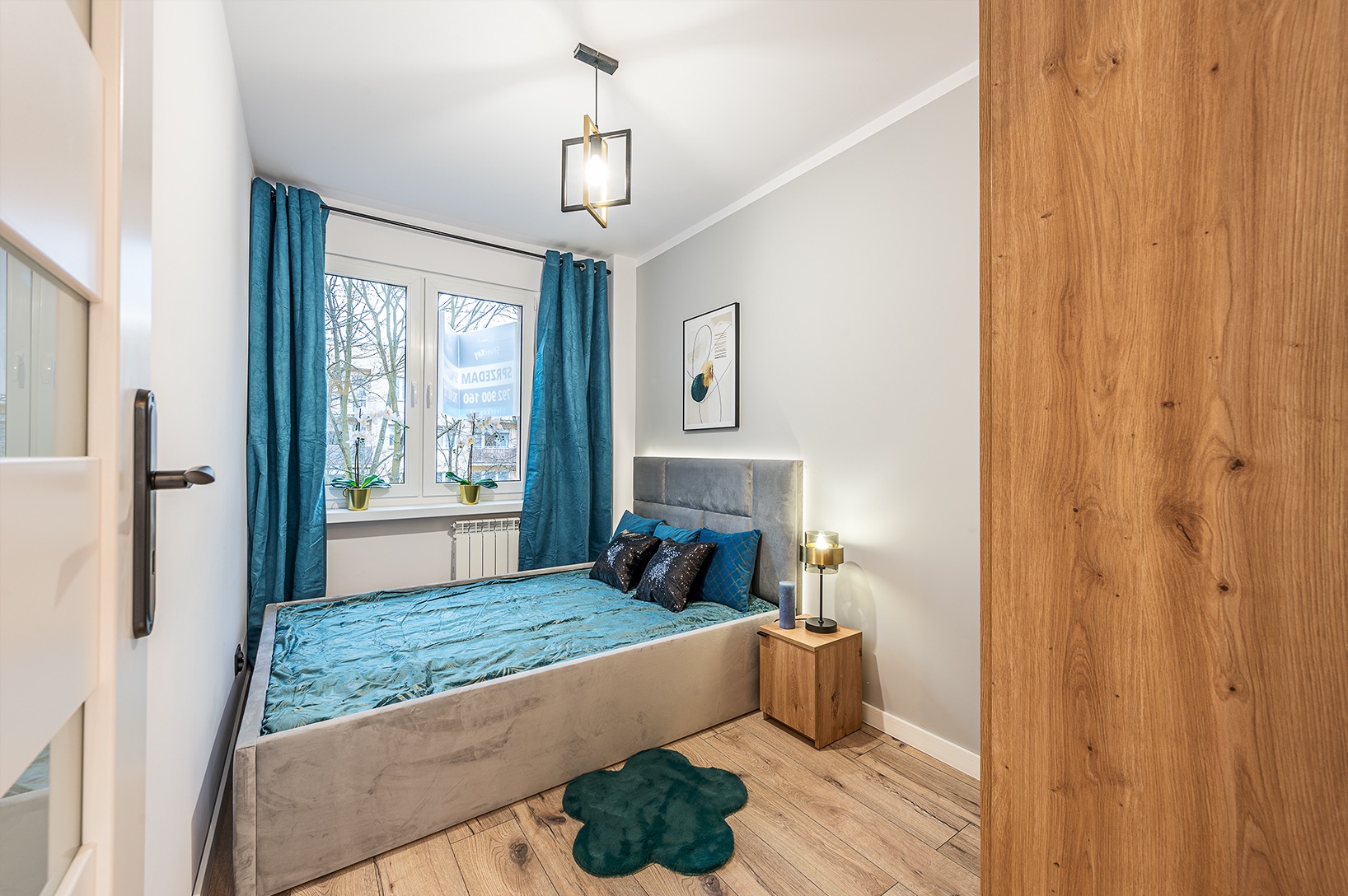 Nowoczesna sypialnia z szarym łóżkiem, turkusowymi zasłonami i drewnianą podłogą. Widoczny fragment drewnianej ściany i lampa na szafce nocnej.