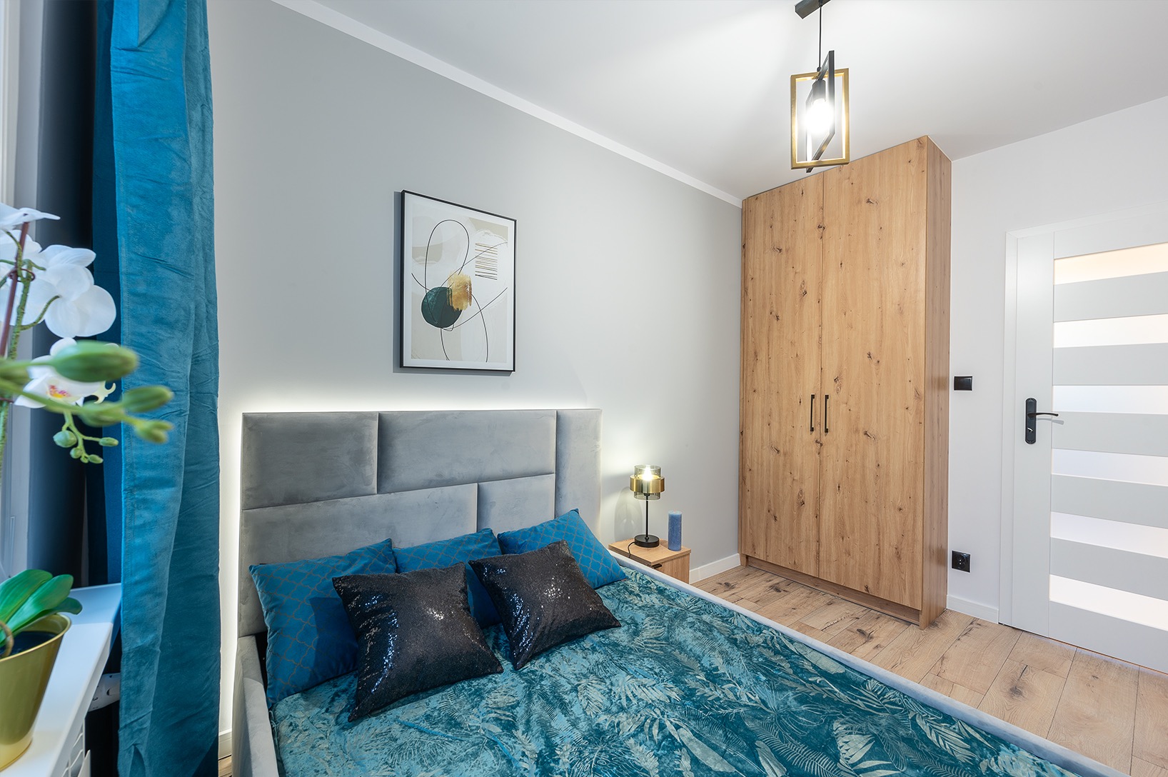 Sypialnia w odcieniach szarości i turkusu: łóżko z podświetlanym wezgłowiem, drewniana szafa, obraz na ścianie i dekoracyjne detale. Nowoczesny design wnętrza.