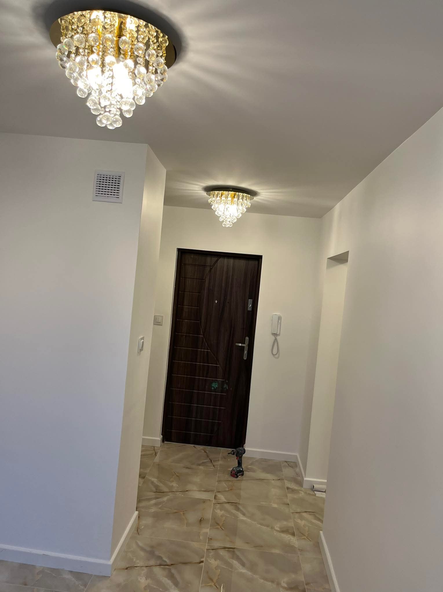 Korytarz po remoncie z dwoma kryształowymi lampami sufitowymi, brązowymi drzwiami i płytkami imitującymi marmur. W rogu widać wiertarkę.