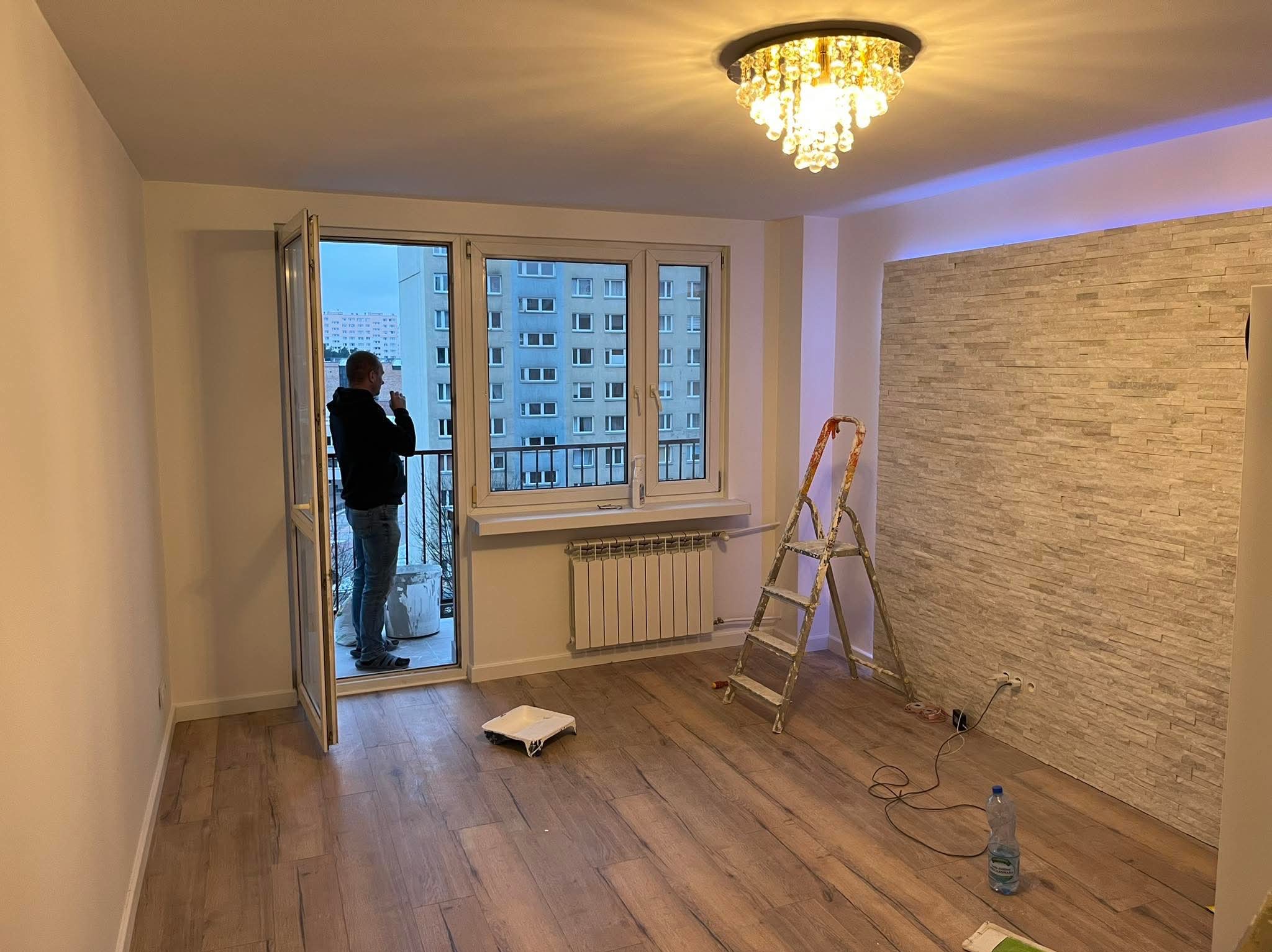 Jasne wnętrze po remoncie: drewnopodobna podłoga, ściana z dekoracyjnej cegły, drabina malarska, otwarte drzwi balkonowe z widokiem na blok. Oświetlenie sufitowe.