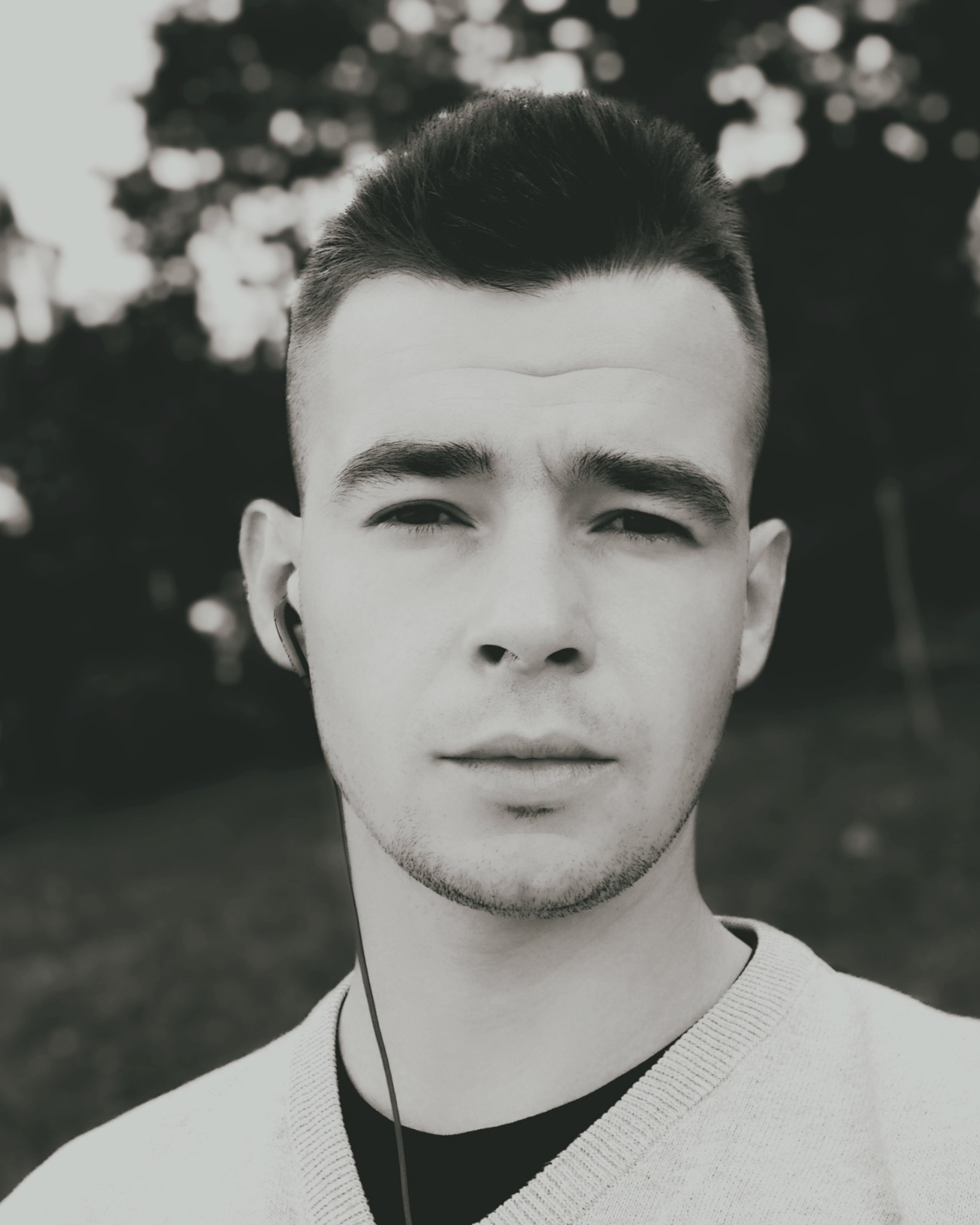 Czarno-biały portret młodego mężczyzny ze słuchawkami dousznymi na tle rozmytego otoczenia. Krótkie włosy, sweter z dekoltem w serek.