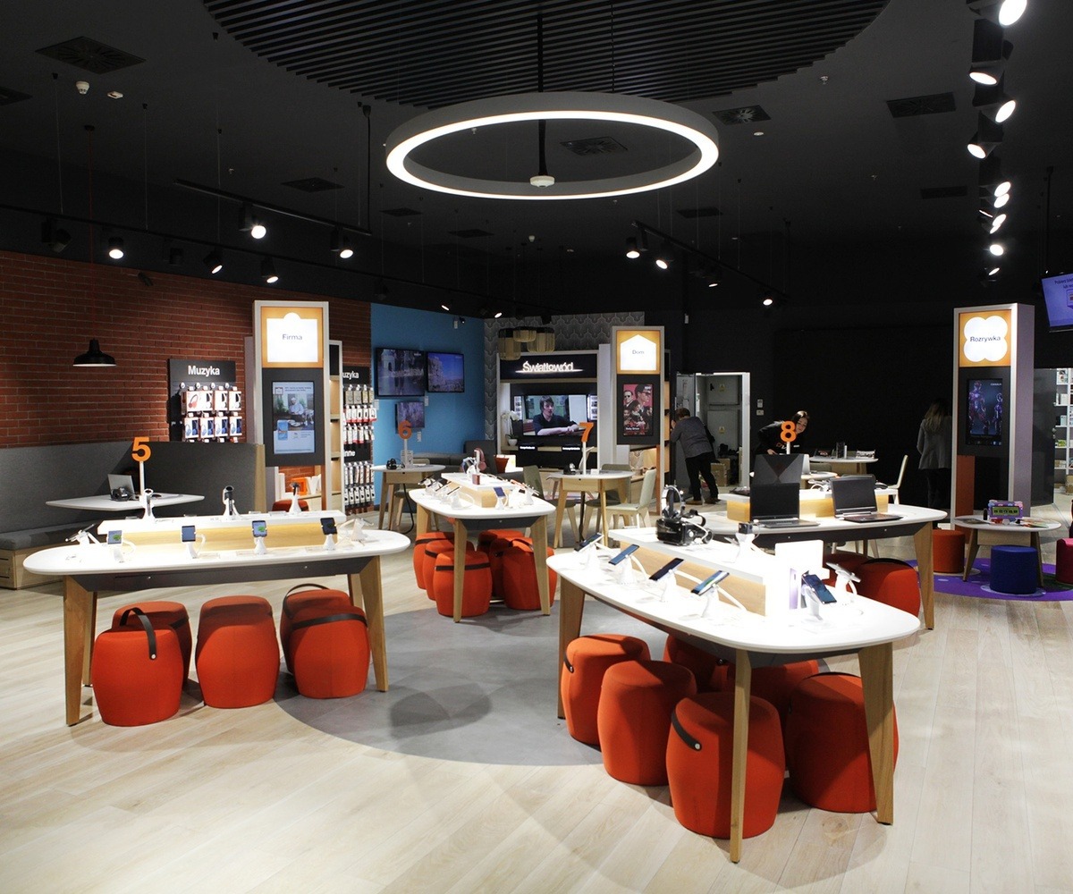 Wnętrze sklepu z elektroniką w Ełku. Ekspozycja smartfonów na białych stołach z pomarańczowymi pufami. Widoczne ekrany reklamowe i oświetlenie punktowe.
