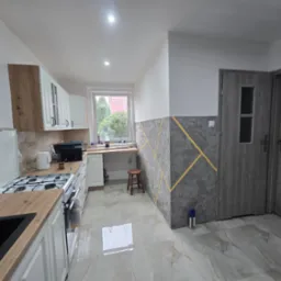 Jasna kuchnia z drewnianym blatem i dekoracyjną ścianą w geometryczne wzory. Widok na okno i drzwi. Błyszczące płytki podłogowe odbijają światło.