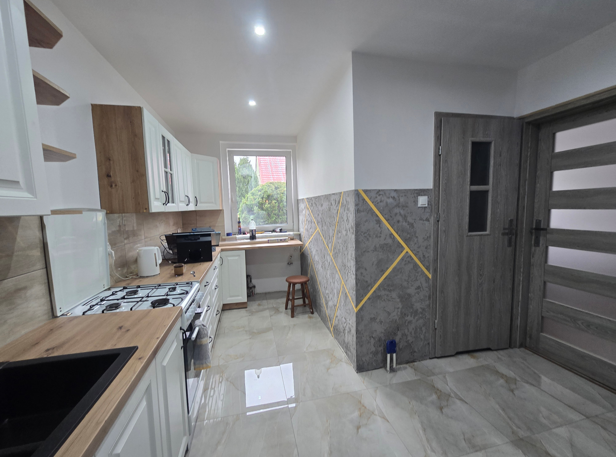 Jasna kuchnia z drewnianym blatem i dekoracyjną ścianą w geometryczne wzory. Widok na okno i drzwi. Błyszczące płytki podłogowe odbijają światło.