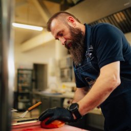 Restauracja & Hotel Złoty Lin - Kucharz z brodą w czarnych rękawicach kroi pomidora na czerwonej desce w profesjonalnej kuchni. Widoczny fartuch z logo i zegarek na ręce.