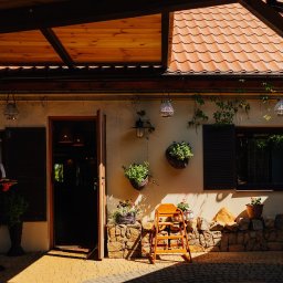 Restauracja & Hotel Złoty Lin - Rustykalna fasada budynku z otwartymi drzwiami, drewnianymi okiennicami i elementami dekoracyjnymi z roślin. W tle widoczny zarys kelnera z tacą.