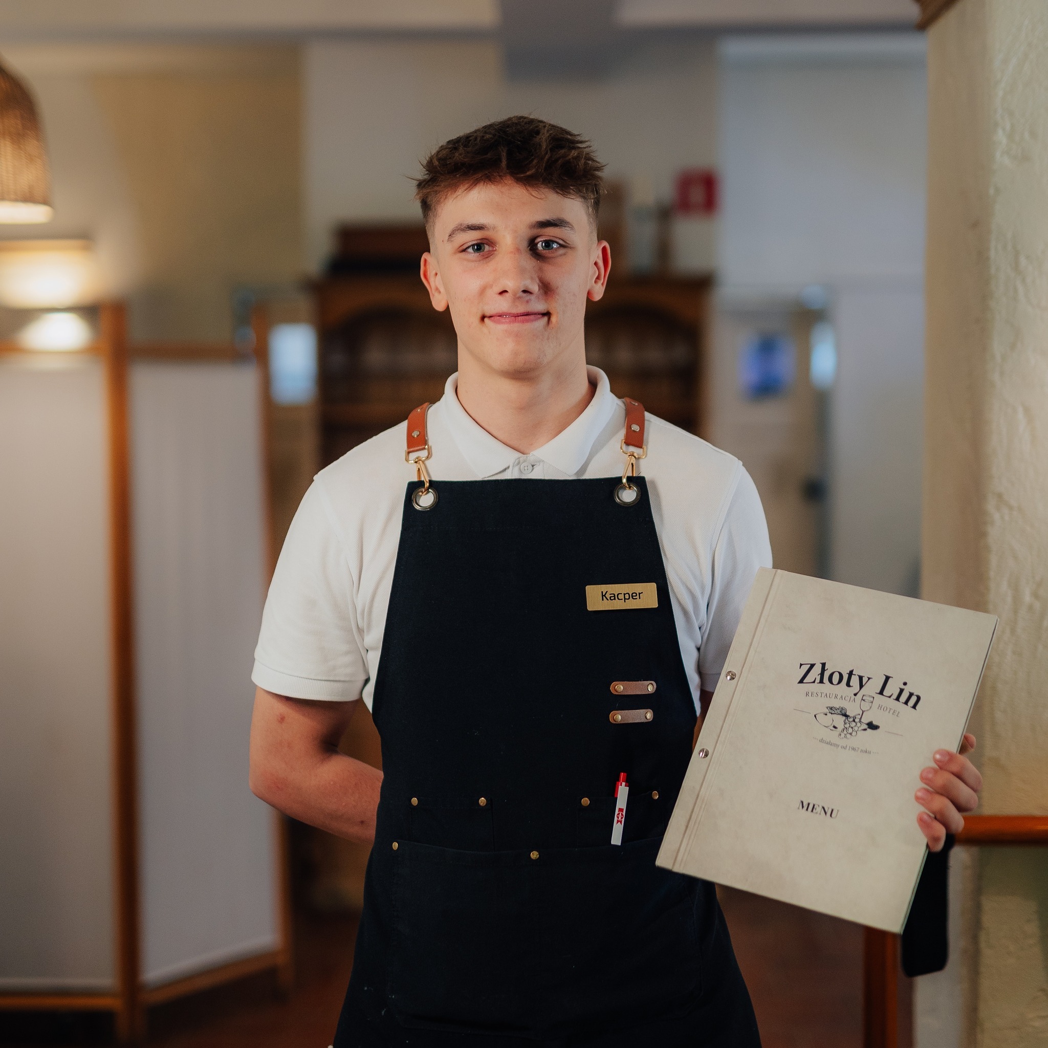 Kelner 'Kacper' w czarnym fartuchu prezentuje menu 'Złoty Lin' w jasnym wnętrzu restauracji. Widoczny fragment baru i dekoracyjne elementy.