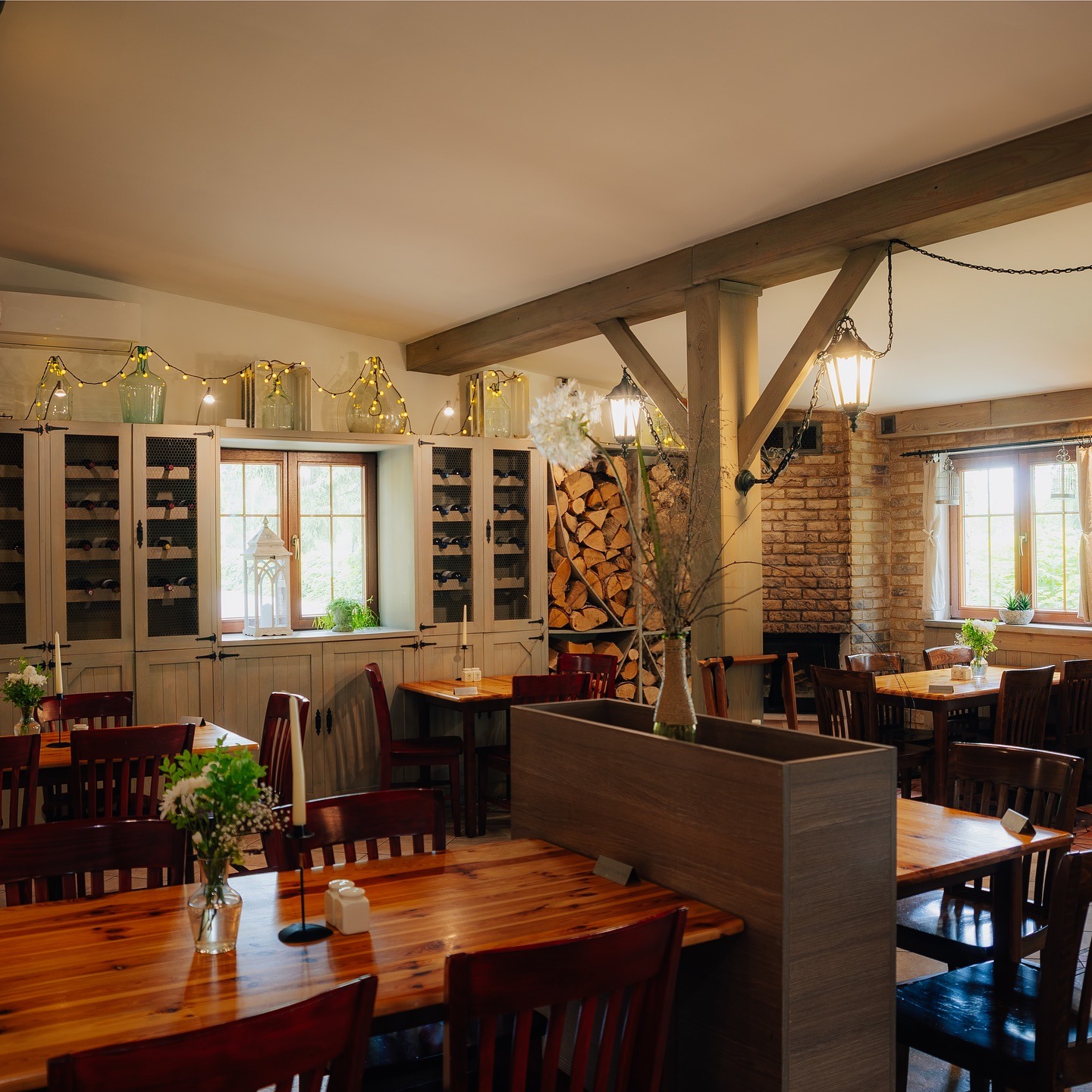Rustykalne wnętrze restauracji z drewnianymi stołami, krzesłami i dekoracjami. W tle kominek z cegły i półki z winami. Ciepłe oświetlenie tworzy przytulną atmosferę.