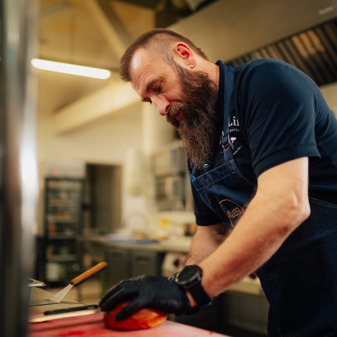 Kucharz z brodą w czarnych rękawicach kroi pomidora na czerwonej desce w profesjonalnej kuchni. Widoczny fartuch z logo i zegarek na ręce.