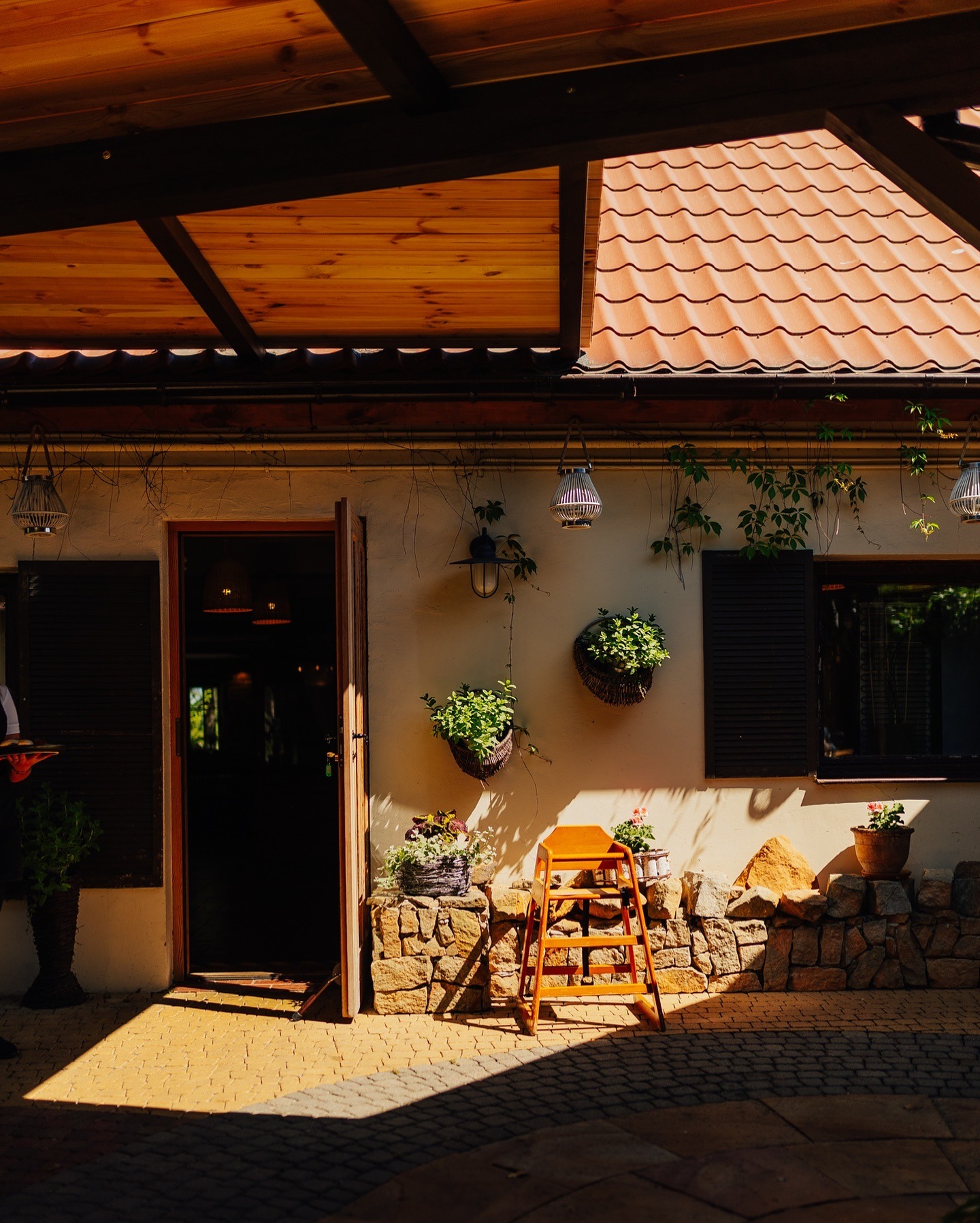 Rustykalna fasada budynku z otwartymi drzwiami, drewnianymi okiennicami i elementami dekoracyjnymi z roślin. W tle widoczny zarys kelnera z tacą.