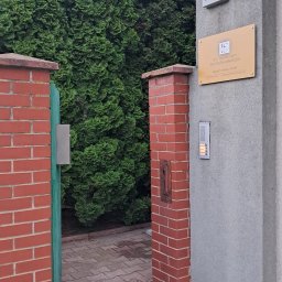 GABINET PSYCHOTRAUMATOLOGICZNY - Wejście do gabinetu z numerem 28, tabliczką informacyjną i domofonem, otoczone zielenią i ceglanymi słupkami.