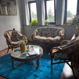 GABINET PSYCHOTRAUMATOLOGICZNY - Przytulny gabinet z rattanowymi meblami w kwiatowe wzory, okrągłym stolikiem kawowym i błękitnym dywanem. W tle trzy okna z widokiem na zieleń.