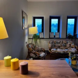 Przytulny gabinet z kwiatowymi fotelami, dyplomami pod oknami i ciepłym oświetleniem lamp. Na pierwszym planie drewniany stół z dekoracyjnymi świecami.