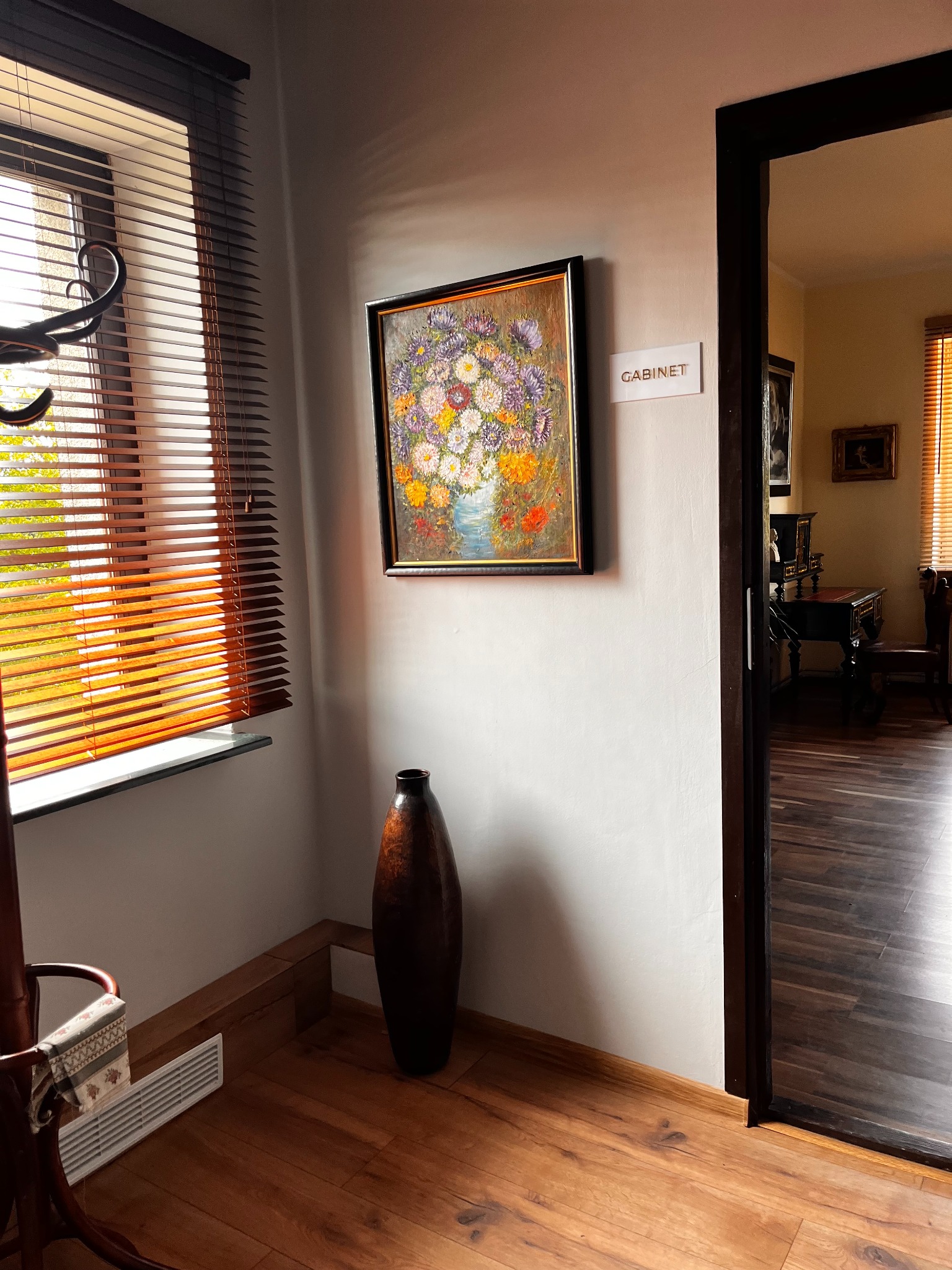 Korytarz z obrazem kwiatów w ramie, brązowym wazonem i widokiem na gabinet przez otwarte drzwi. Żaluzje w oknie wpuszczają ciepłe światło.