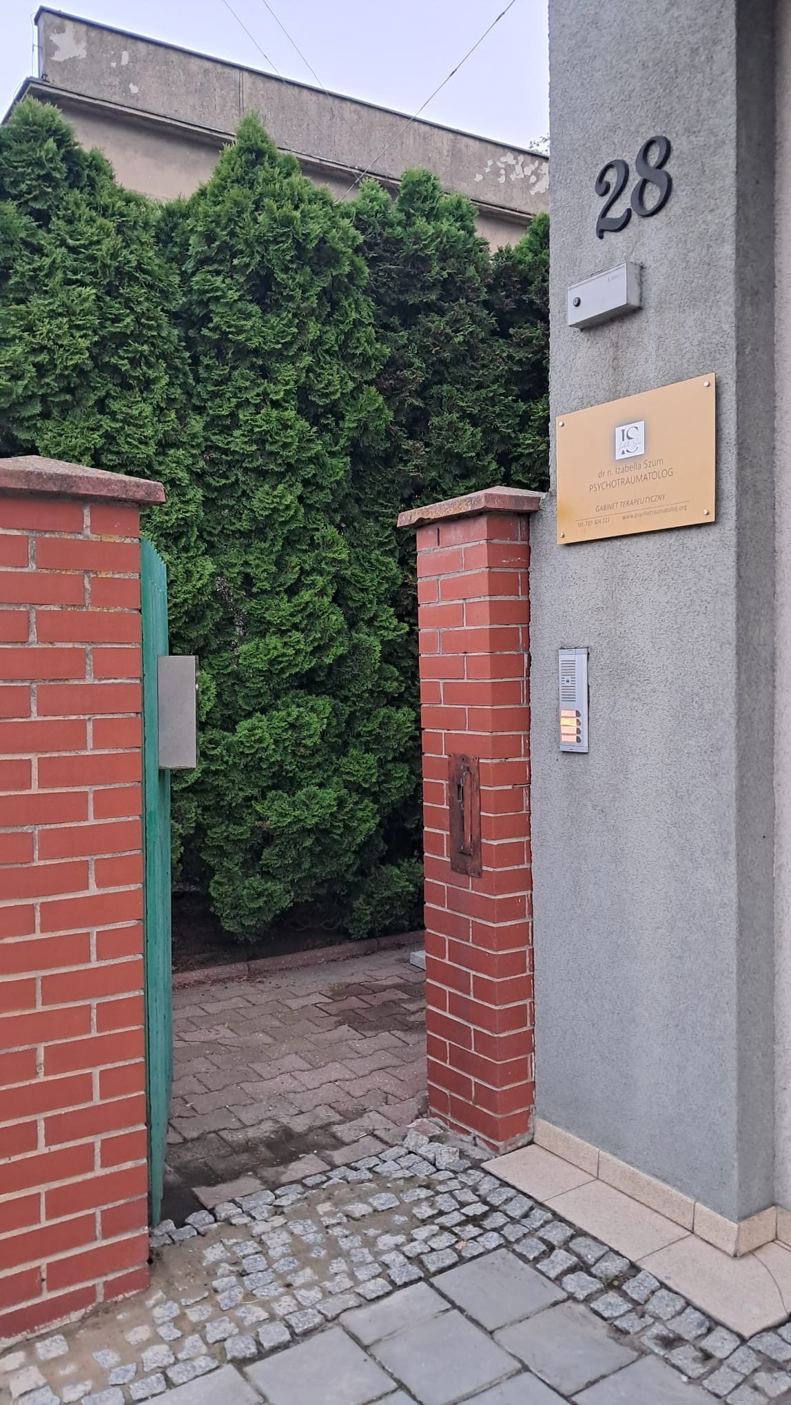 Wejście do gabinetu z numerem 28, tabliczką informacyjną i domofonem, otoczone zielenią i ceglanymi słupkami.