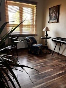 Kącik relaksacyjny: fotel w kwiaty, pianino, lampka na stoliku, obraz na ścianie. Przytulne wnętrze z drewnianą podłogą i widokiem z okna.