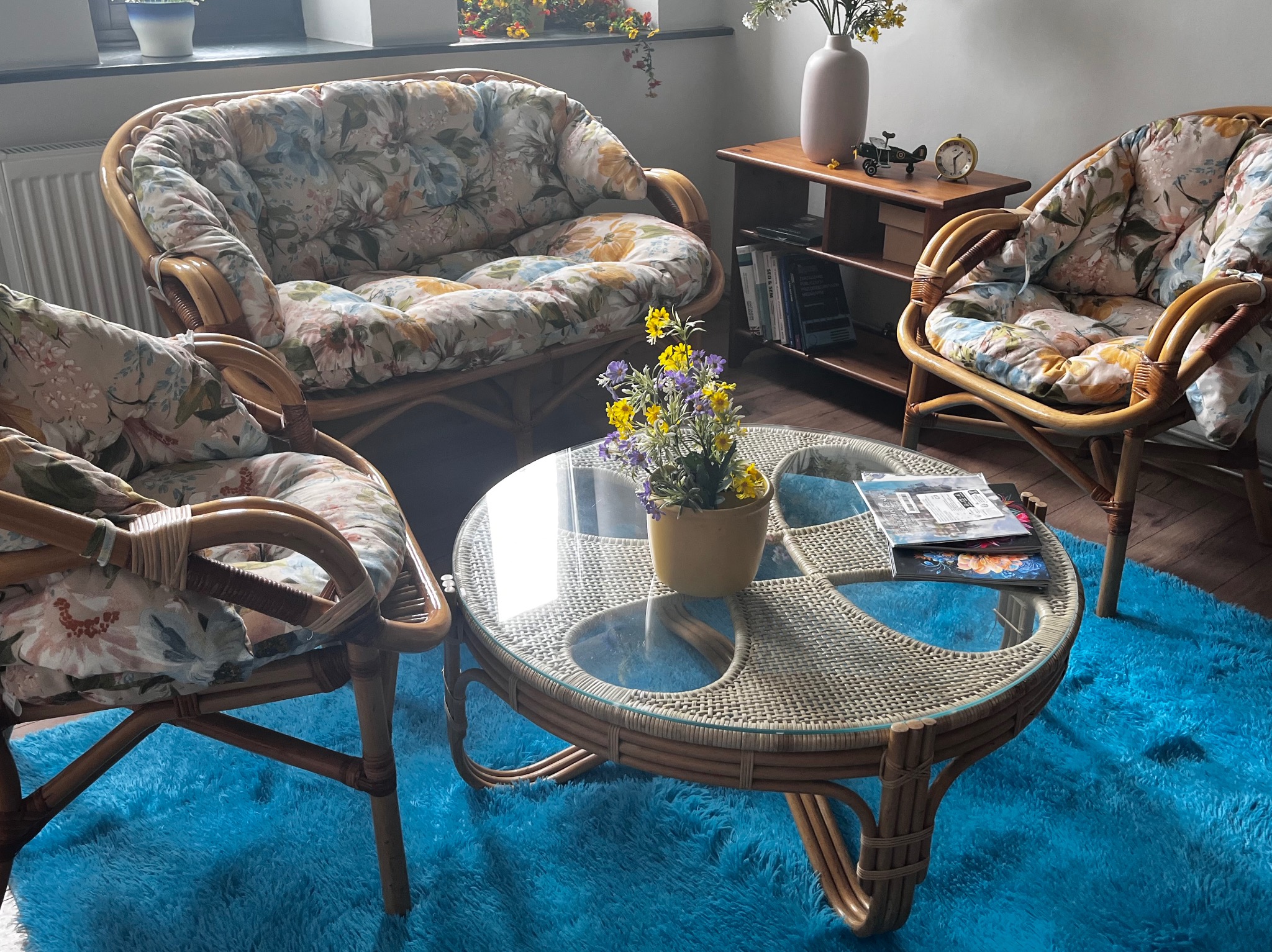 Przytulny kącik relaksacyjny z rattanowymi meblami w kwiatowe wzory i turkusowym dywanem, tworzący spokojną atmosferę w jasnym wnętrzu.