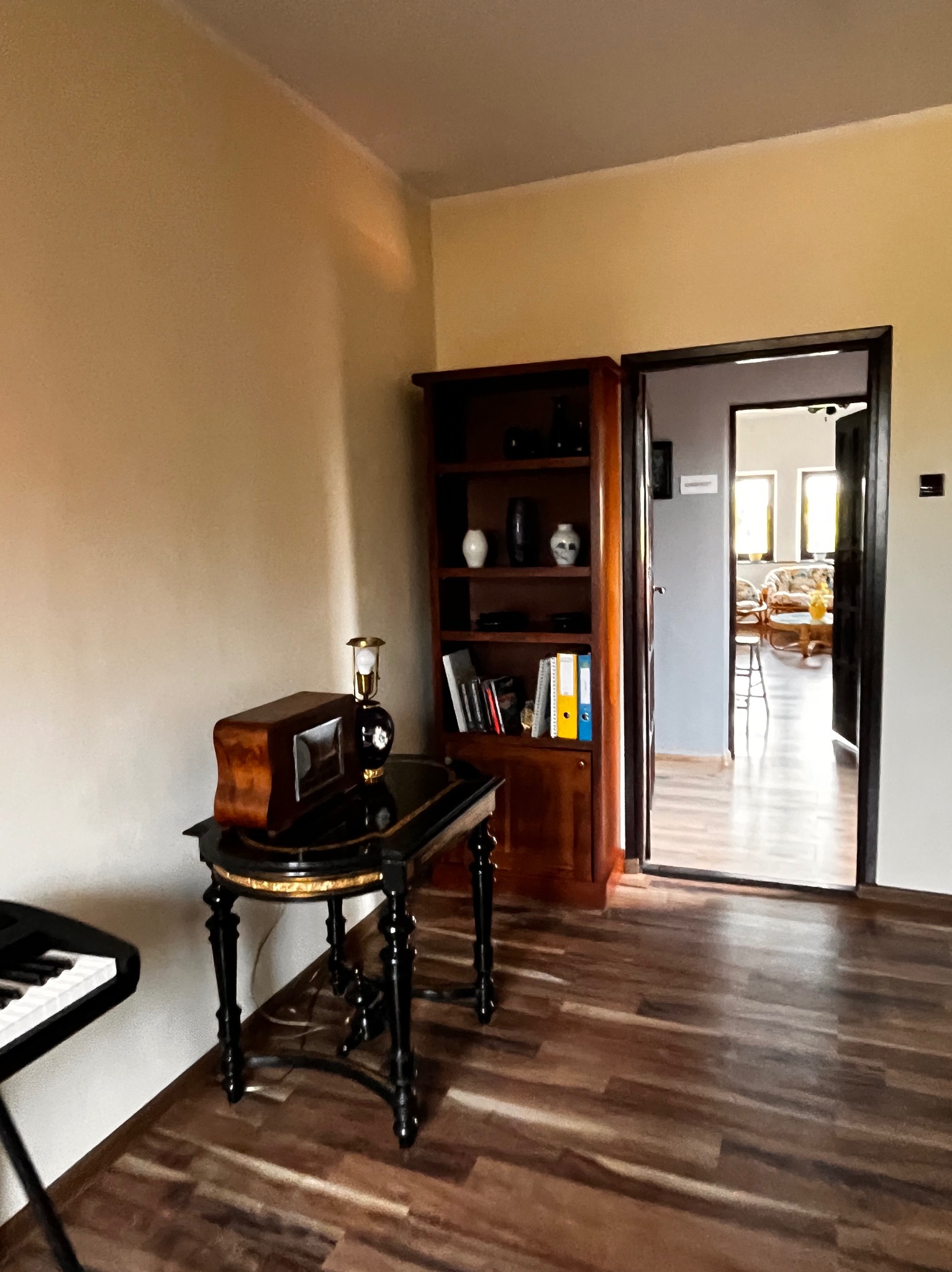 Ustawny gabinet z pianinem, regałem i lampką retro na stoliku. W tle widok na jasny pokój dzienny przez otwarte drzwi, tworzący przyjazną atmosferę.