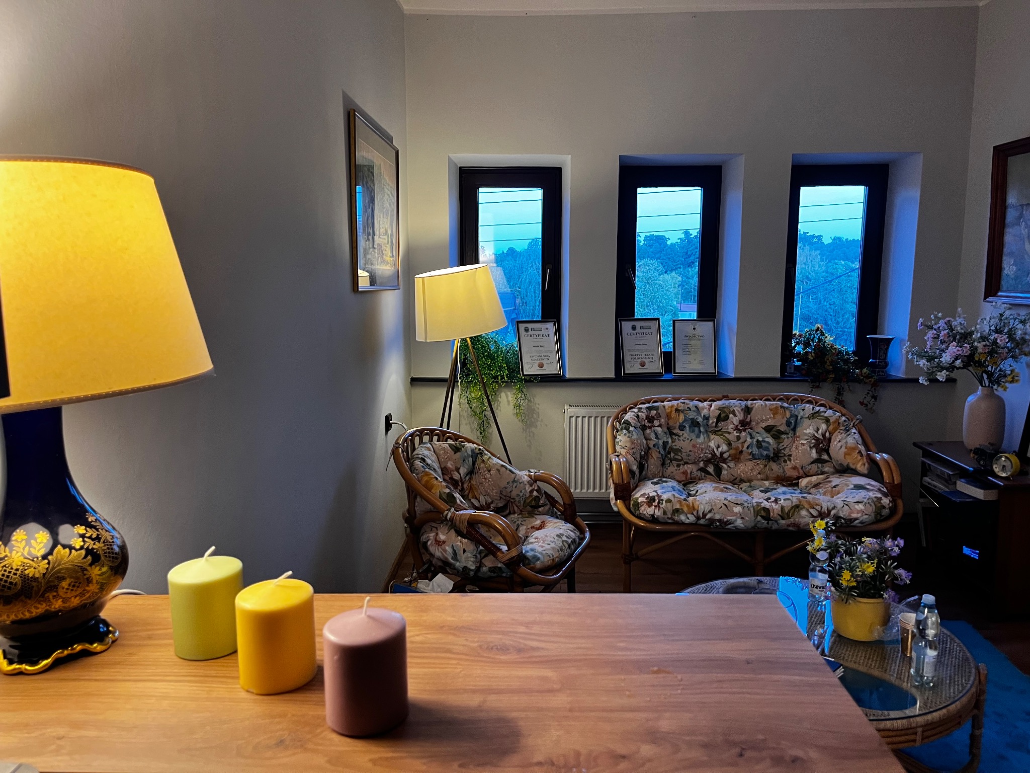 Przytulny gabinet z kwiatowymi fotelami, dyplomami pod oknami i ciepłym oświetleniem lamp. Na pierwszym planie drewniany stół z dekoracyjnymi świecami.