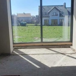 TEST-POL MARDEUSZ I ŻAK SPÓŁKA JAWNA - Wnętrze surowego domu z dużym oknem tarasowym, przez które widać domy i zielony trawnik. Na podłodze widoczne fragmenty instalacji.