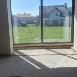 Wnętrze surowego domu z dużym oknem tarasowym, przez które widać domy i zielony trawnik. Na podłodze widoczne fragmenty instalacji.