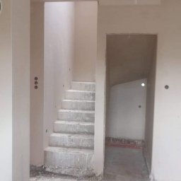 TEST-POL MARDEUSZ I ŻAK SPÓŁKA JAWNA - Surowe schody betonowe w jasnym wnętrzu budynku w stanie deweloperskim. Widoczne ślady po pracach budowlanych i instalacje elektryczne. Otwór drzwiowy w tle.