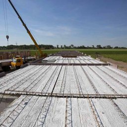 TEST-POL MARDEUSZ I ŻAK SPÓŁKA JAWNA - Montaż stropu z prefabrykatów betonowych na budowie domu jednorodzinnego. Widoczny żuraw, ciężarówka i zielone pola w tle.