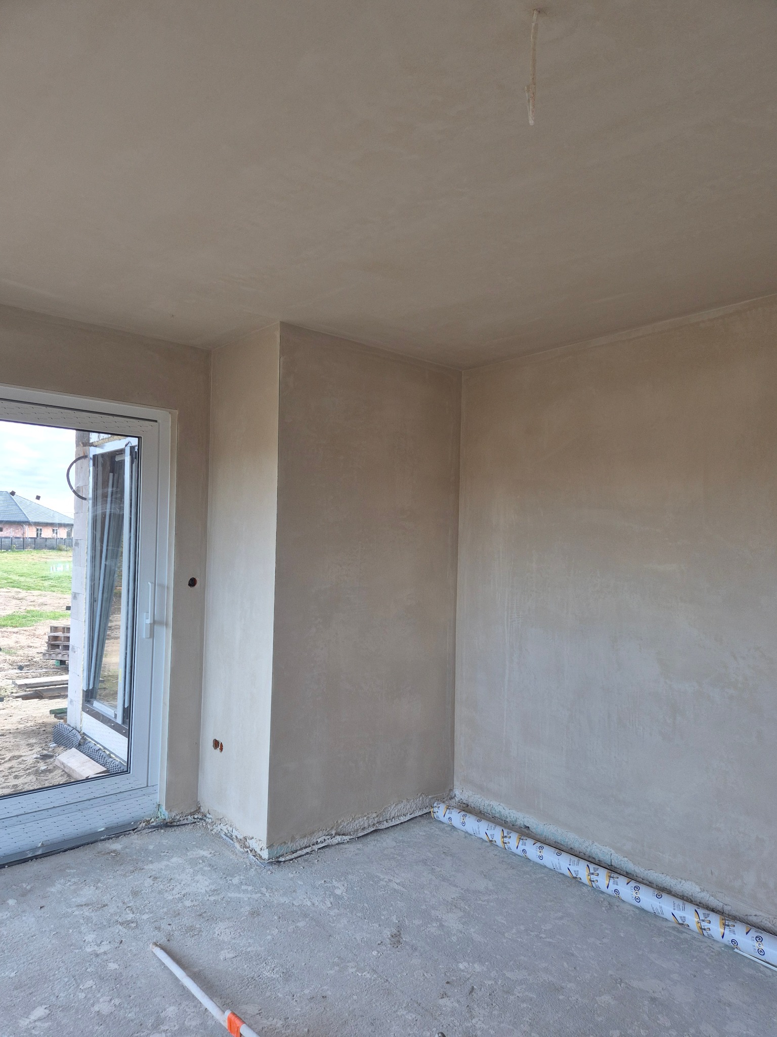 Wnętrze po tynkowaniu maszynowym: gładkie, jasne ściany w surowym pomieszczeniu z oknem i widocznymi przewodami. Przy podłodze rolka folii zabezpieczającej.