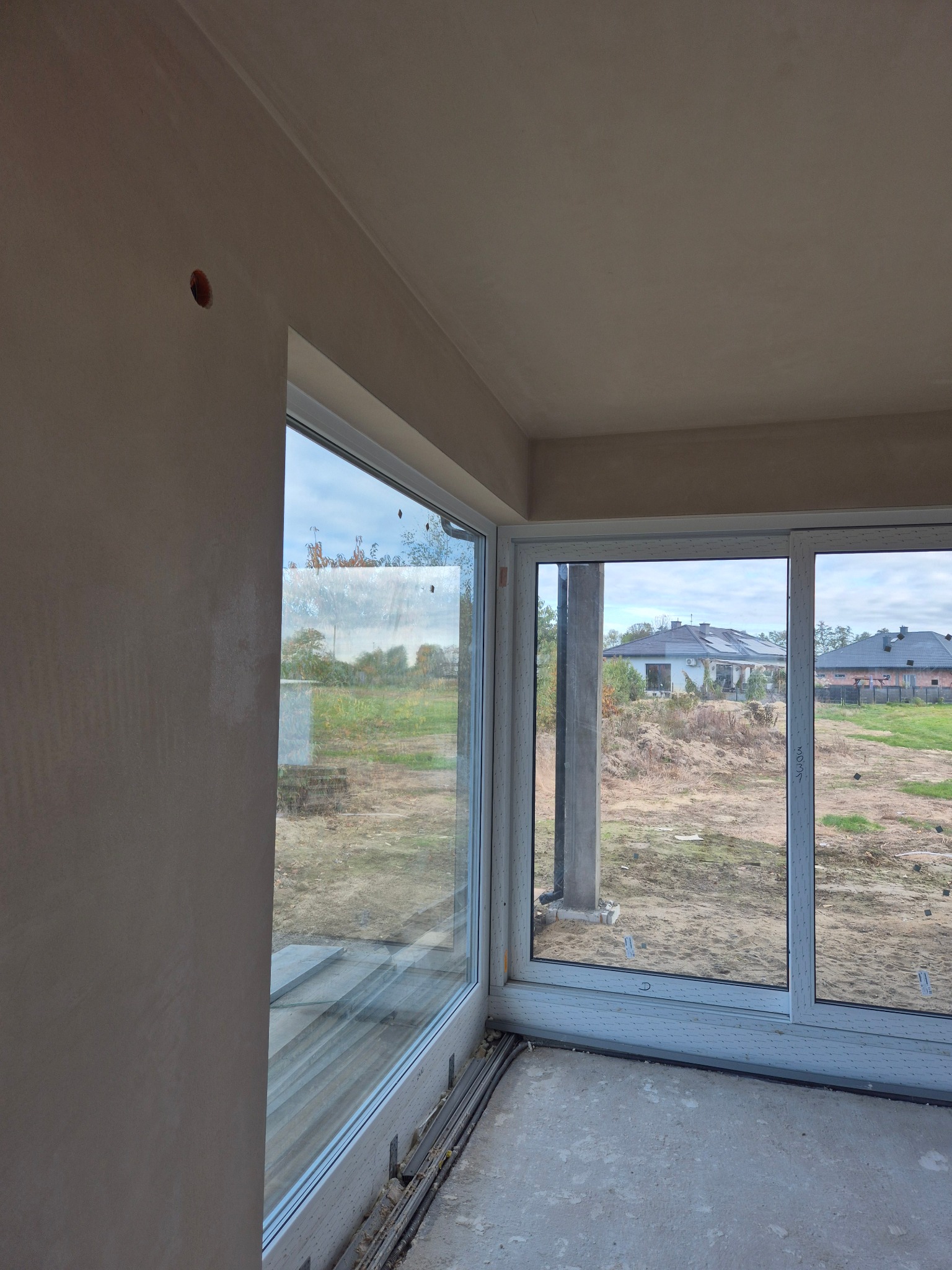 Wnętrze w trakcie wykańczania: świeżo otynkowana ściana z widocznym otworem na instalację, duże okna z widokiem na domy i niezagospodarowany teren.