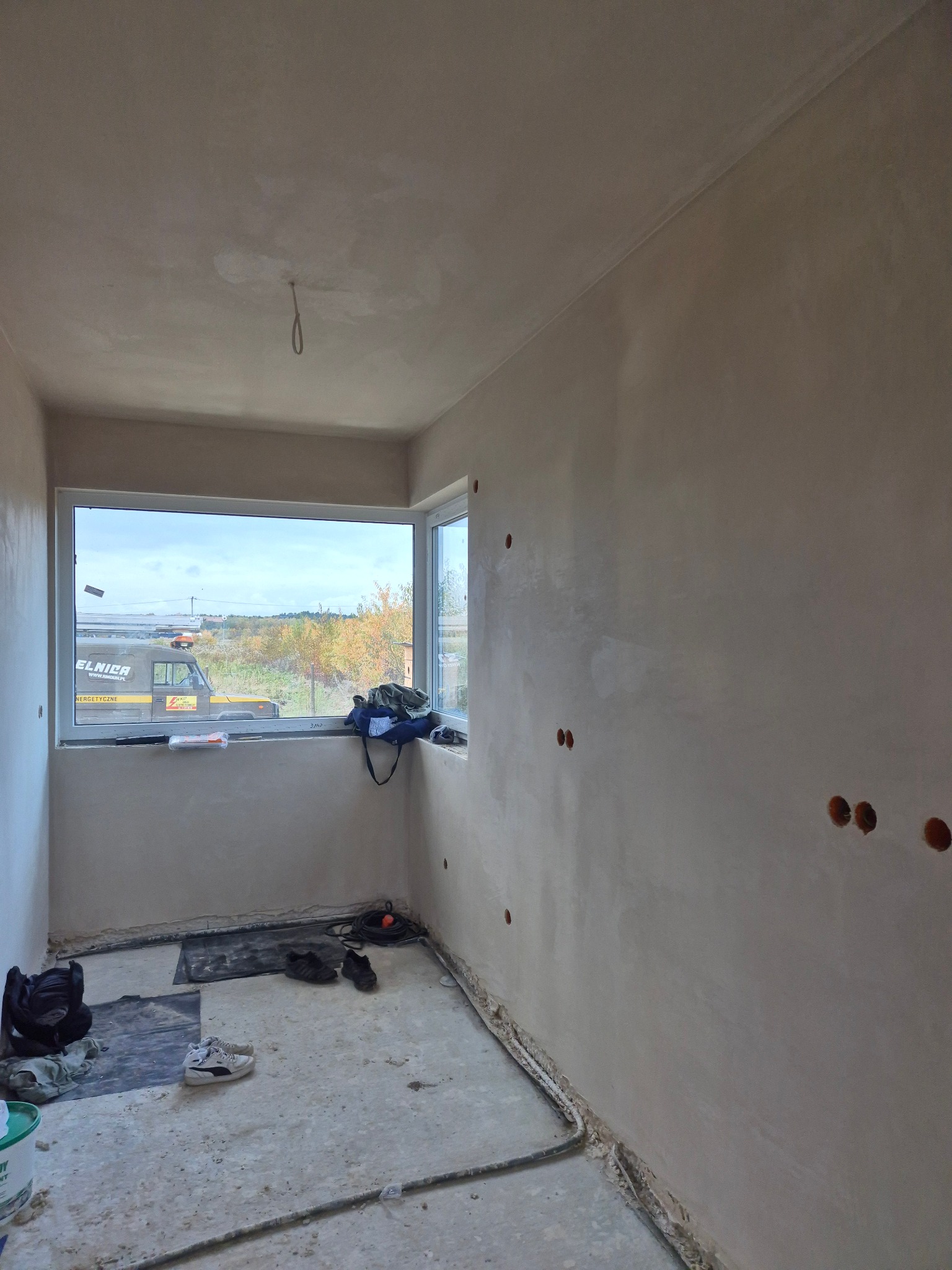 Pokój po tynkowaniu maszynowym ścian i sufitu, widoczne otwory na gniazdka elektryczne, okno z widokiem na zewnątrz. Na podłodze leżą narzędzia i buty.