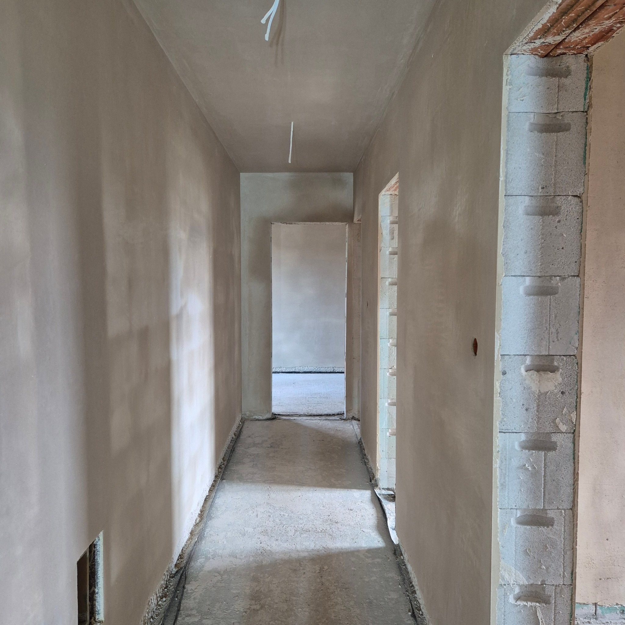 Korytarz w trakcie wykańczania: ściany pokryte świeżym, jasnym tynkiem, widoczne bloczki betonowe przy wejściu do pomieszczenia, surowy beton na podłodze.