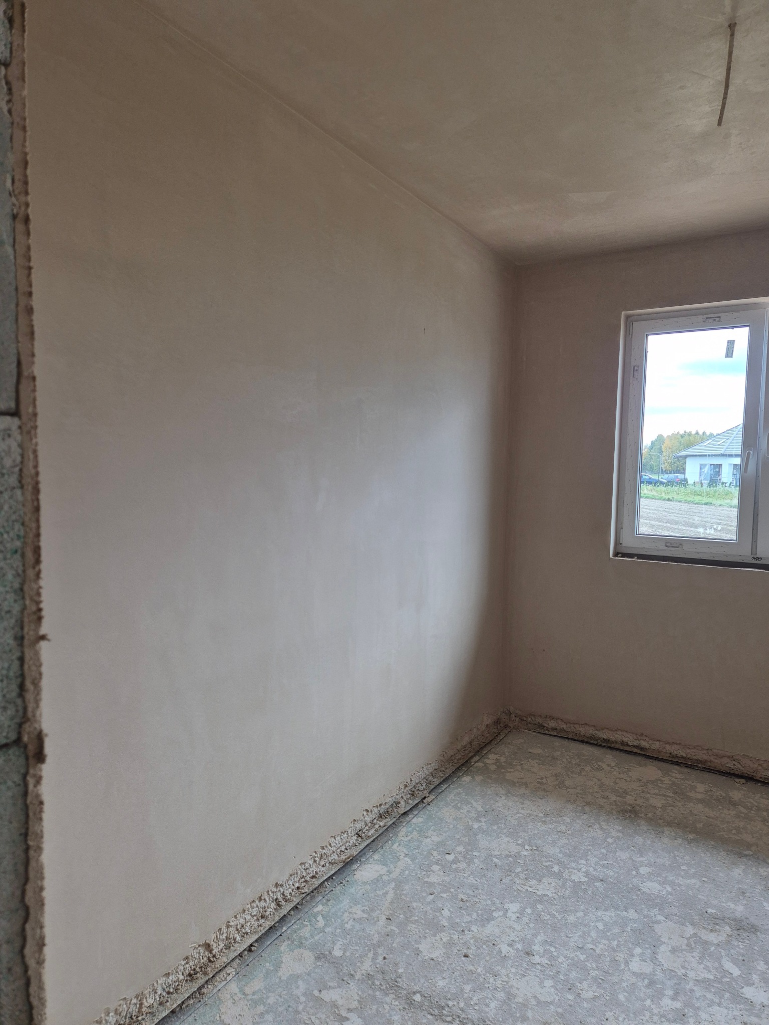 Wnętrze pokoju po nałożeniu gładzi szpachlowej na ściany i sufit. Widoczne okno z widokiem na zewnątrz oraz surowa podłoga. Wykończone ściany mają jasny, beżowy odcień.