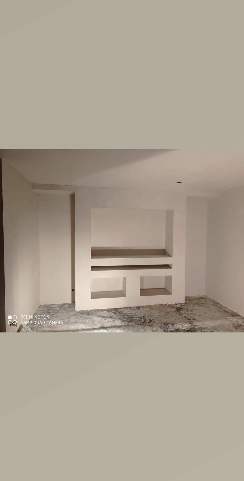 Biała wnęka ścienna z półkami w surowym, jasnym wnętrzu. Widoczne ślady po pracach wykończeniowych na podłodze. Minimalistyczny design, brak dekoracji.