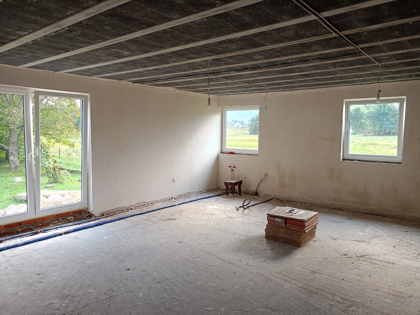 Surowy stan wnętrza domu z betonowym stropem, trzema oknami i przygotowaną instalacją podłogową. Na podłodze leżą płytki, a w oknach widoczne drzewa.