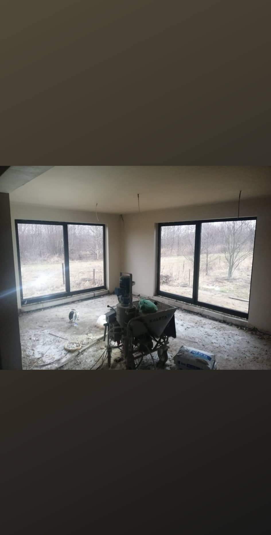 Wnętrze w trakcie prac tynkarskich: maszyna do tynkowania, worek z zaprawą i widok na zewnątrz przez duże okna. Ściany przygotowane do dalszych prac wykończeniowych.