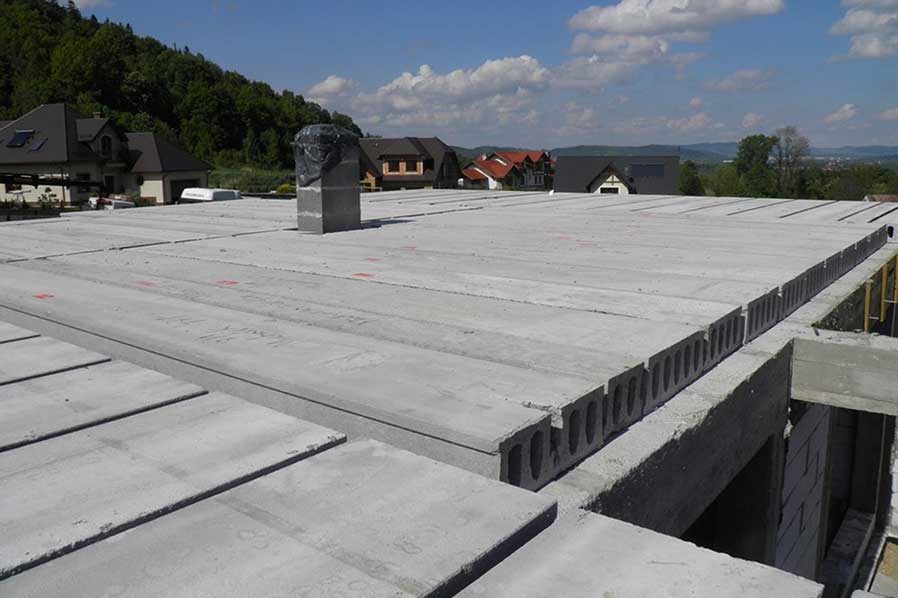 Szare, prefabrykowane płyty stropowe z otworami, ułożone na budowie domu, z widocznym kominem i zabudową w tle, w słoneczny dzień.