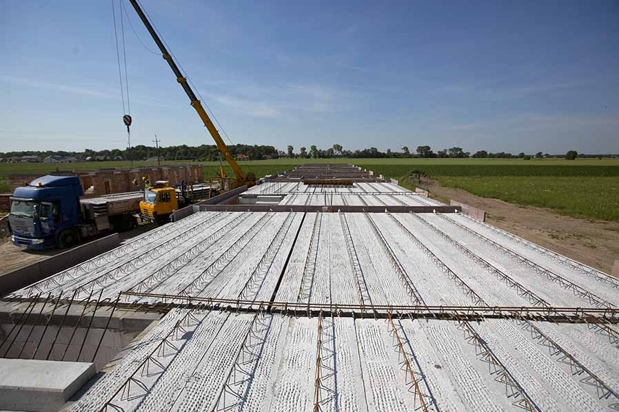 Montaż stropu z prefabrykatów betonowych na budowie domu jednorodzinnego. Widoczny żuraw, ciężarówka i zielone pola w tle.