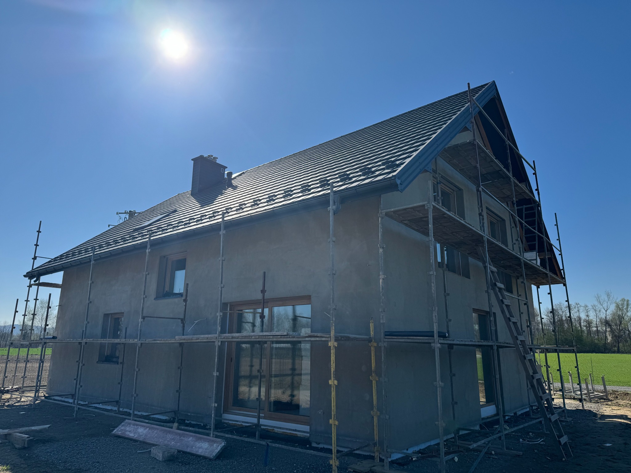 Surowy dom jednorodzinny w trakcie tynkowania, otoczony rusztowaniami, z widocznym dachem z dachówki i oknami. Słoneczny dzień, błękitne niebo.